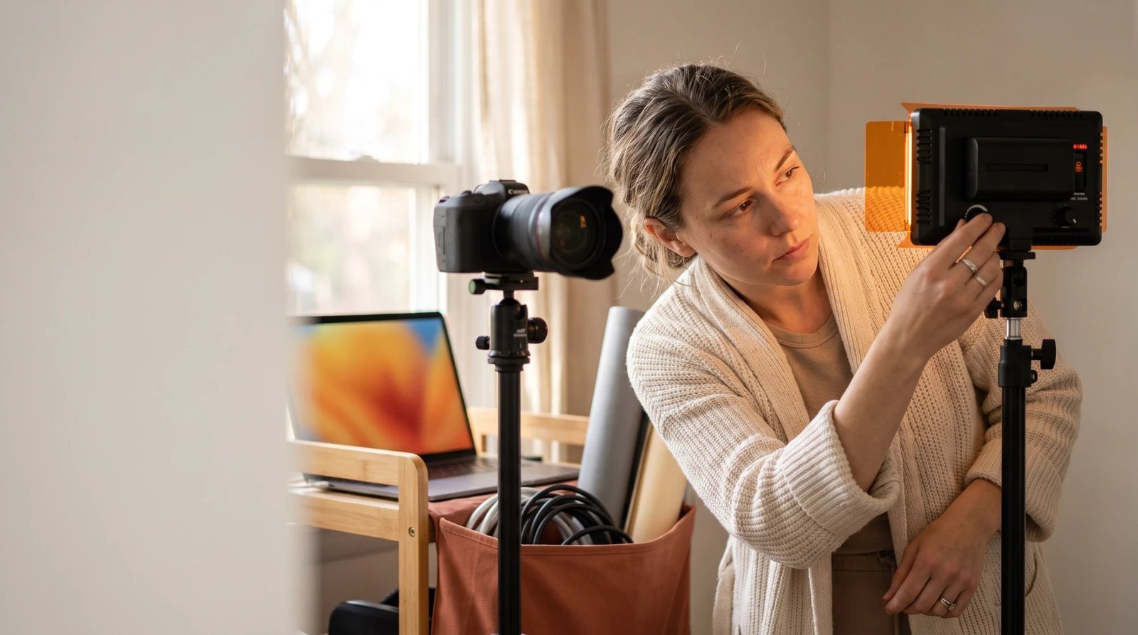 A woman in a cream cardigan adjusting the dial on an LED video light with an orange gel next to a camera on a tripod, with a laptop and a bag of cables on a wooden cart by a window